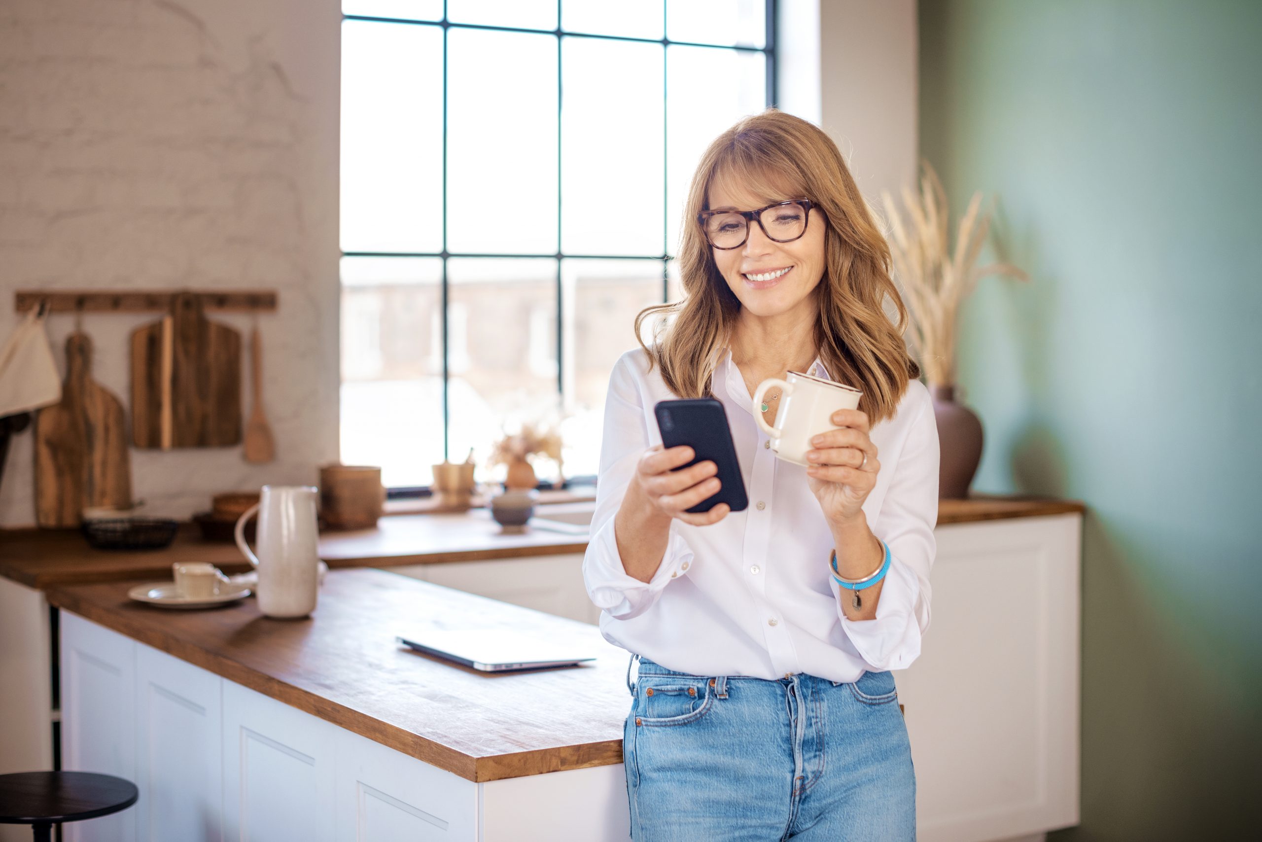 Smiling woman looking at cellphone and drinking coffee while standing at her kitchen in the morning