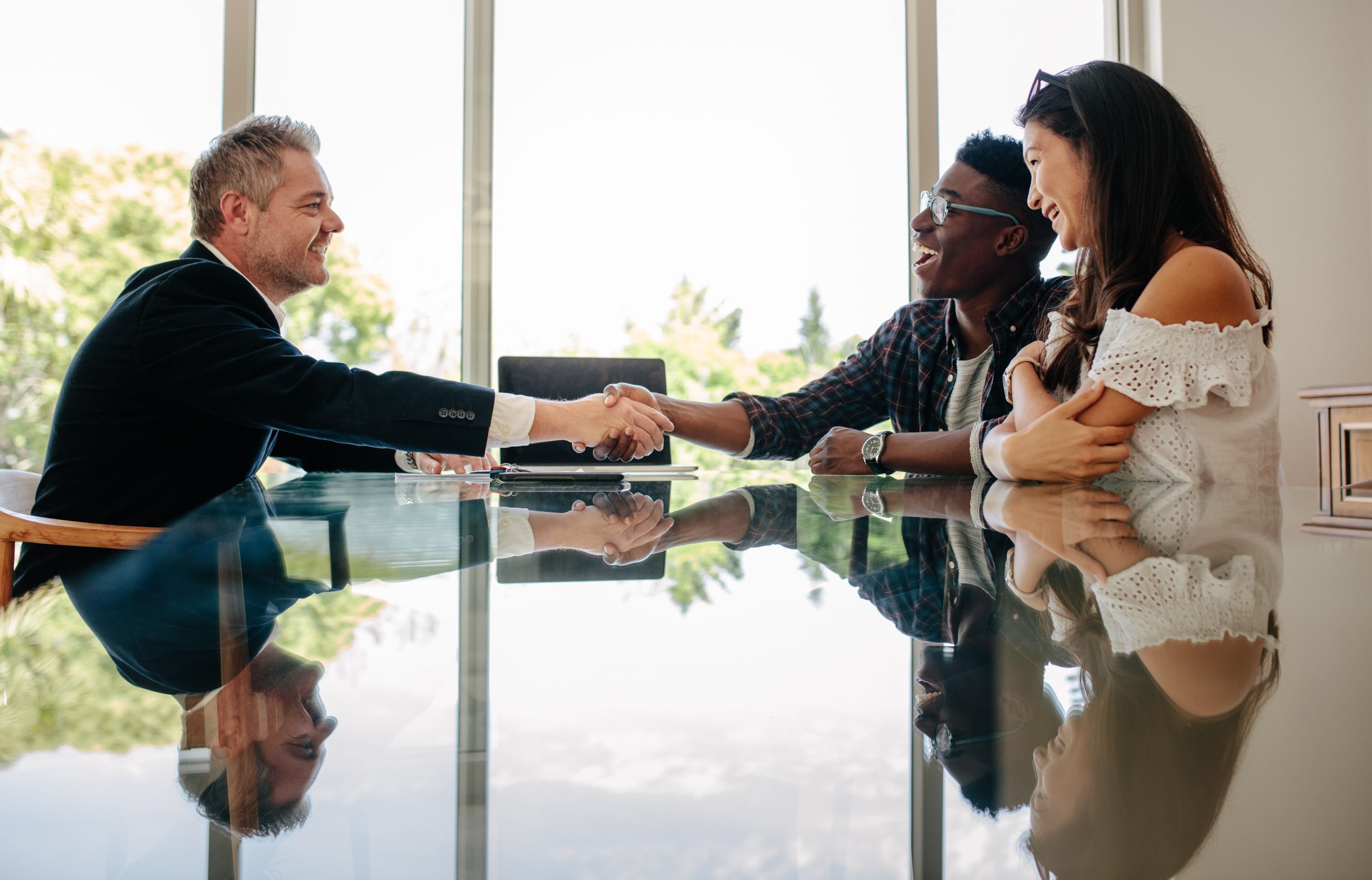 Male real estate broker shaking hands with new property owners while sitting across a table. Property seller congratulating couple on making deal on new house.