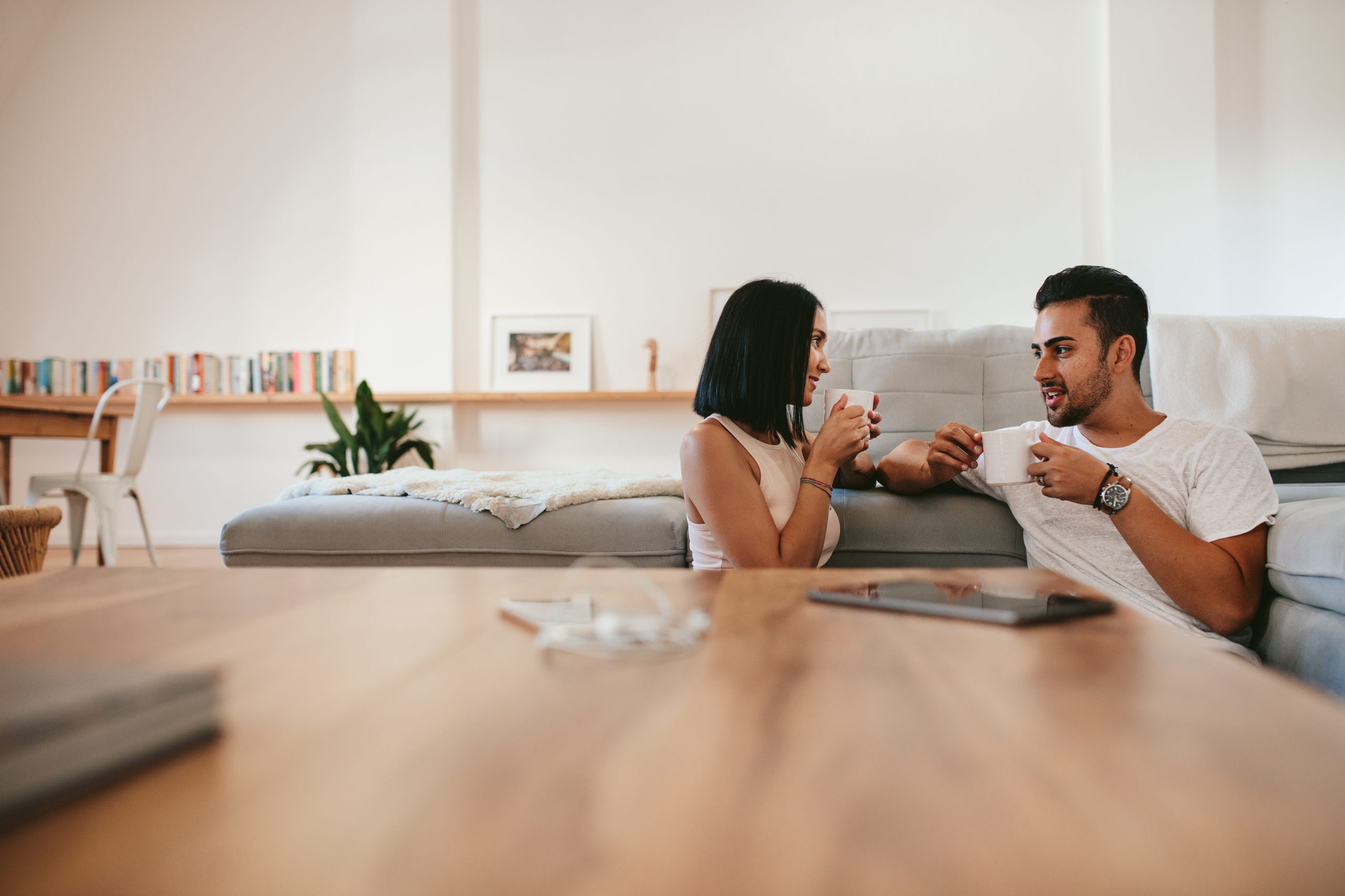 Young couple relaxing by the couch with cup of coffee in the living room. Man and woman sitting together at home having coffee and talking.